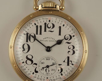 May include: A gold pocket watch with a white face and black hands. The watch has a second hand and a smaller dial for seconds. The watch face has the text "HAMILTON RAILWAY SPECIAL".