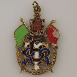 May include: A gold-colored pendant with a red, white, and blue enamel design. The pendant features a crown with a red and white enamel design, two flags, and the letters "K.R." in blue enamel.