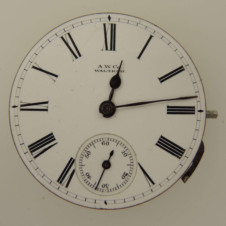 May include: A white pocket watch face with black Roman numerals and hands. The watch face has a smaller subdial at the bottom and the text "AWC Co. WALTHAM" is printed at the top.