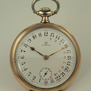 May include: A gold-toned Omega pocket watch with a white face and black numerals. The watch features a subdial and a silver-colored loop at the top. The watch hands are gold-toned.