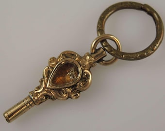 May include: A gold-toned pocket watch key with an ornate design and a clear glass insert. The key has a ring attached to it.