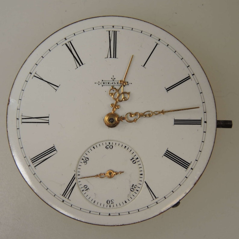 May include: A white pocket watch with Roman numeral hour markers and black hands. The watch has a smaller, circular sub-dial with black markings and a gold hand. The watch face is inscribed with the word "ELGIN".
