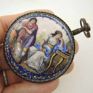 May include: Antique, round, hand-painted enamel compact with a detailed scene of two figures in classical attire. The compact has a silver-toned metal frame and a winding key. The artwork features a blue and floral border.