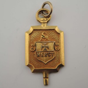 May include: A gold-colored pendant with a shield design. The pendant features a textured surface and a smaller shield with a cross and the word "MERCY." It has a loop at the top for hanging.