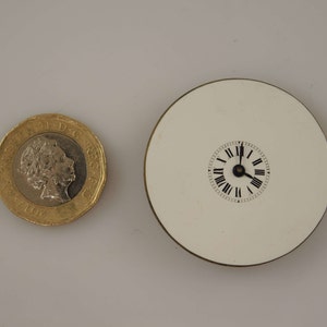May include: A white circular clock face with black Roman numeral markings and black hands. The clock is mounted on a white circular base with a gold rim.