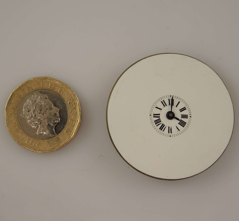 May include: A white circular clock face with black Roman numeral markings and black hands. The clock is mounted on a white circular base with a gold rim.