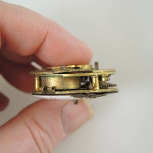 K&ouml;nnte beinhalten: Nahaufnahme einer zerlegten antiken Taschenuhr von der Seite mit vergoldeten Zahnr&auml;dern und einem wei&szlig;en Zifferblatt.
