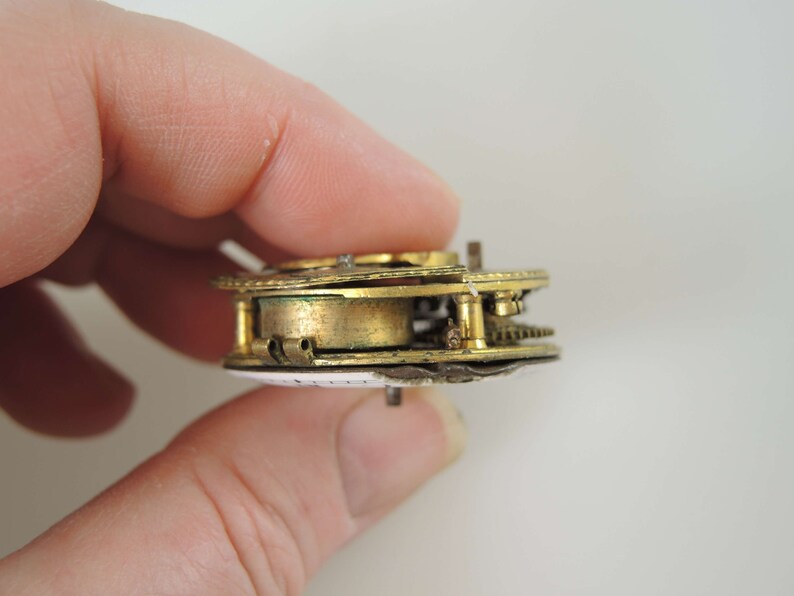 K&ouml;nnte beinhalten: Nahaufnahme einer zerlegten antiken Taschenuhr von der Seite mit vergoldeten Zahnr&auml;dern und einem wei&szlig;en Zifferblatt.