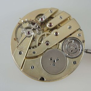 Puede incluir: Primer plano de un movimiento de reloj de bolsillo de tono dorado con engranajes intrincados y un v&aacute;stago de cuerda. El movimiento es visible a trav&eacute;s de una tapa de vidrio transparente.