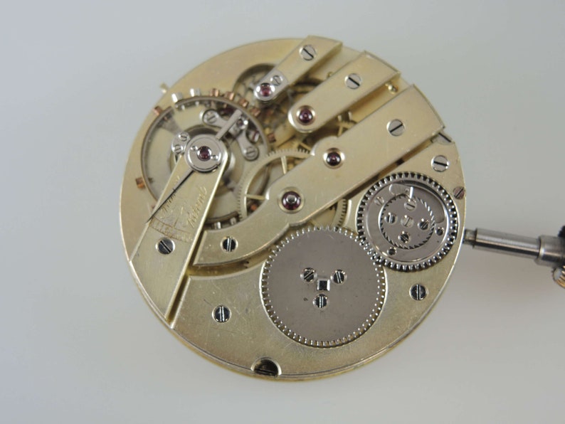 Puede incluir: Primer plano de un movimiento de reloj de bolsillo de tono dorado con engranajes intrincados y un v&aacute;stago de cuerda. El movimiento es visible a trav&eacute;s de una tapa de vidrio transparente.
