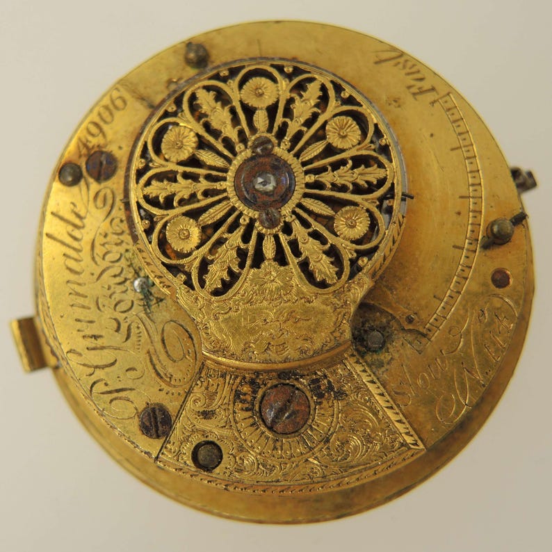 Puede incluir: Primer plano del movimiento de un reloj de bolsillo de oro vintage. El mecanismo del reloj presenta intrincados filigranas florales, texto grabado y peque&ntilde;os tornillos. La esfera del reloj es de color oro c&aacute;lido, con una peque&ntilde;a piedra en forma de diamante en el centro.