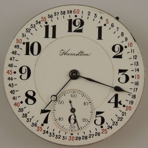 Puede incluir: Un reloj de bolsillo blanco con marcadores de hora en números romanos negros y agujas negras. La esfera del reloj tiene un círculo concéntrico más pequeño con marcadores de minutos en números arábigos negros y una aguja negra. La esfera del reloj también tiene la palabra "Hamilton" impresa en escritura negra. La esfera del reloj está rodeada por un anillo negro con números rojos.