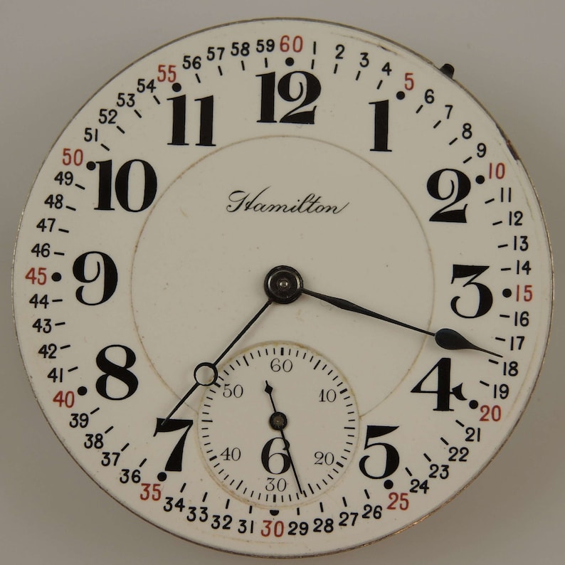 May include: A white pocket watch with black Roman numeral hour markers and black hands. The watch face has a smaller, concentric circle with black Arabic numeral minute markers and a black hand. The watch face also has the word "Hamilton" printed in black script. The watch face is surrounded by a black ring with red numbers.