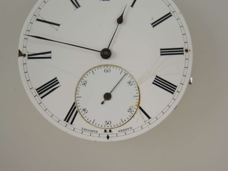 Op de afbeelding: Een wit zakhorloge-wijzerplaat met zwarte Romeinse cijfers voor de uren en zwarte markeringen voor de minuten. Het horloge heeft een kleinere, witte sub-wijzerplaat met zwarte markeringen voor de minuten. De wijzerplaat van het horloge is gesigneerd met "Lecomte Geneve".