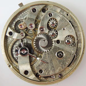 May include: Detailed view of a vintage pocket watch mechanism. The silver-coloured gears, springs, and levers are visible, with ruby jewels and engraved details. The watch's inner workings are displayed in detail.