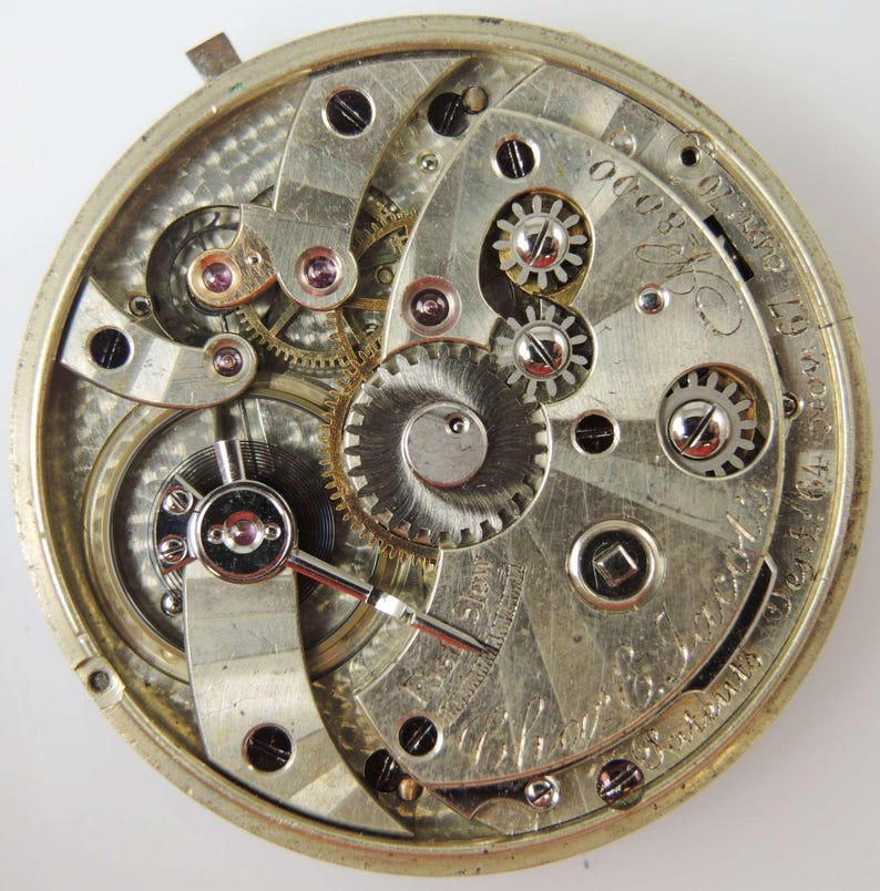 May include: Detailed view of a vintage pocket watch mechanism. The silver-coloured gears, springs, and levers are visible, with ruby jewels and engraved details. The watch's inner workings are displayed in detail.