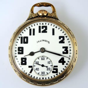May include: A gold-tone pocket watch with a white dial, black Arabic numerals, and the word ILLINOIS printed in black. It features a small sub-dial for seconds at the bottom, black hands, and an ornate, engraved gold-tone bow at the top crown.