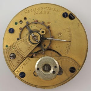 Puede incluir: Primer plano de un movimiento de reloj antiguo. El mecanismo dorado presenta intrincados engranajes, tornillos y un volante. El texto "SPRINGFIELD, MASS." es visible en la parte superior, con el n&uacute;mero "317285" y "PATENT PINION".
