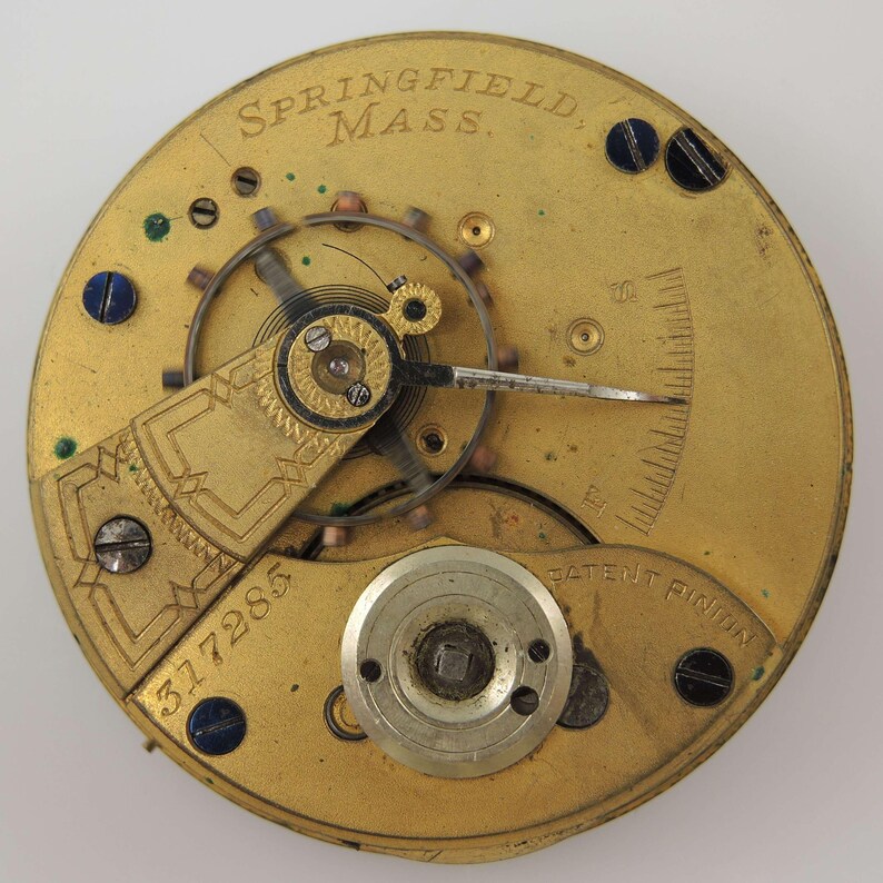 Puede incluir: Primer plano de un movimiento de reloj antiguo. El mecanismo dorado presenta intrincados engranajes, tornillos y un volante. El texto "SPRINGFIELD, MASS." es visible en la parte superior, con el n&uacute;mero "317285" y "PATENT PINION".