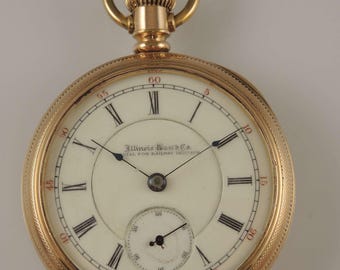 Puede incluir: Un reloj de bolsillo de oro con números romanos en la esfera. El reloj tiene un pequeño subdial y el texto "Illinois Watch Co. Ideal for Railway Service" está impreso en la esfera.