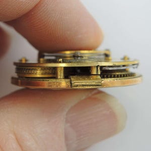 Puede incluir: Primer plano de un movimiento de reloj de bolsillo de tono dorado con engranajes intrincados y una caja de lat&oacute;n.
