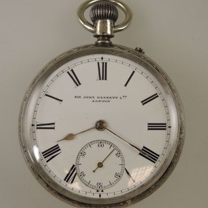 May include: A silver pocket watch with a white face and black Roman numerals. The watch face includes a smaller seconds dial and the text "SIR JOHN BENNETT LTD LONDON". The watch has a decorative silver ring at the top.