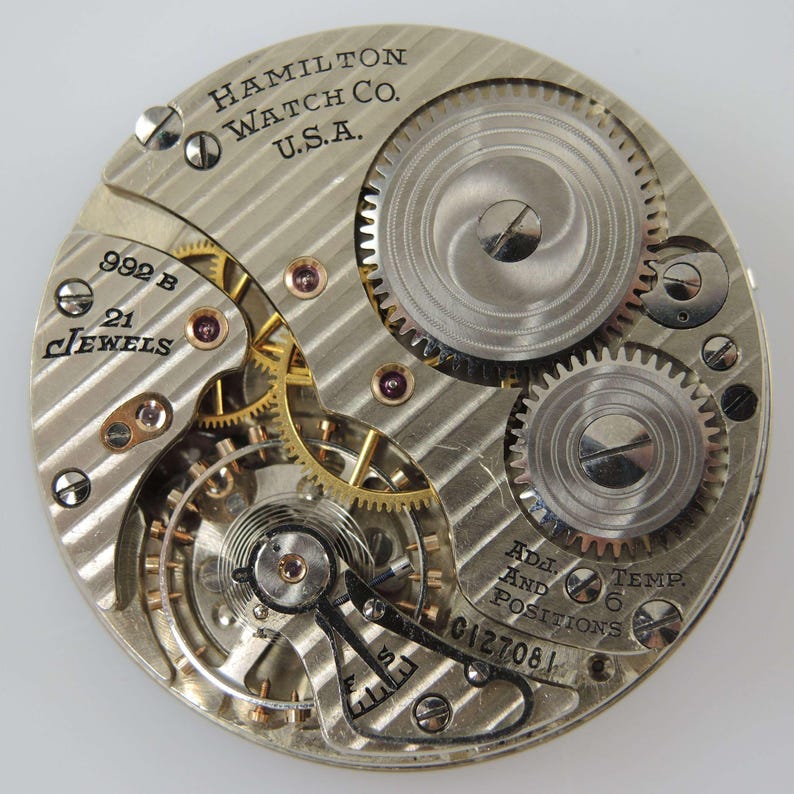Pu&ograve; includere: Primo piano del movimento di un orologio Hamilton Watch Co. U.S.A. Il meccanismo argentato presenta ingranaggi intricati, gioielli e il testo "992 B 21 Jewels". Il testo aggiuntivo include "Adj. And Temp. 6 Positions" e "0127081".