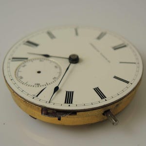 May include: A white pocket watch face with black Roman numeral hour markers and black hands. The watch has a small, white sub-dial with black markings.