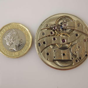 May include: Close-up of a gold-toned watch movement with intricate gears and a silver-toned plate. The movement is surrounded by a circular frame with a small coin for scale.