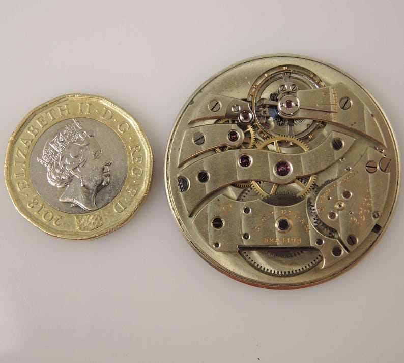 May include: Close-up of a gold-toned watch movement with intricate gears and a silver-toned plate. The movement is surrounded by a circular frame with a small coin for scale.