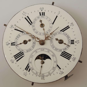 May include: A white clock face with Roman numeral hour markers and black markings for the minutes. The clock has three smaller dials for the day of the week, the month, and the moon phase. The clock face is surrounded by a white metal frame.