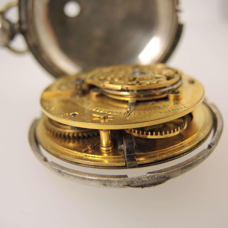 May include: Close-up of an open antique pocket watch, revealing the intricate gold-colored gears and mechanisms. The watch casing is silver, and the internal components are meticulously detailed, showcasing the craftsmanship of the timepiece.