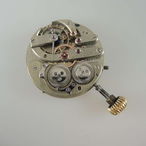 May include: A silver watch movement with intricate gears and a gold winding stem. The movement is exposed, showing the inner workings of the watch.