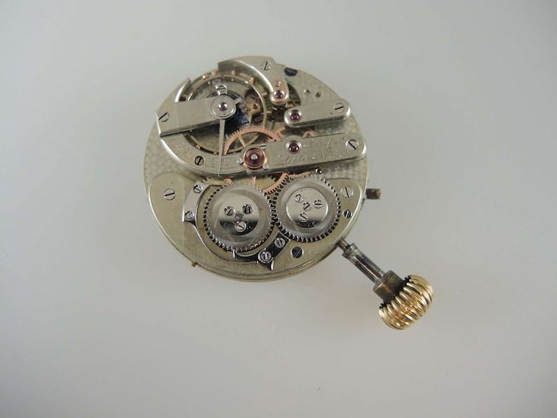 May include: A silver watch movement with intricate gears and a gold winding stem. The movement is exposed, showing the inner workings of the watch.