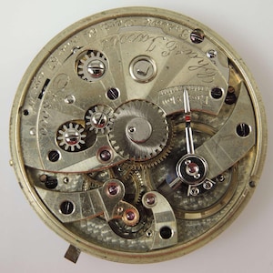 May include: Detailed view of a vintage watch mechanism, showcasing a complex arrangement of gears, springs, and other components. The silver-coloured metal parts are arranged in a circular design, with visible text and markings. A fine example of precision engineering.