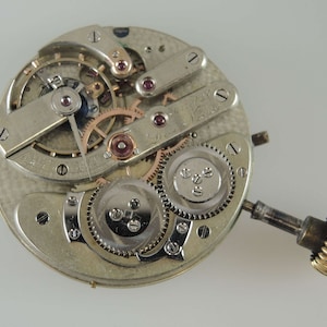 May include: Close-up of a silver pocket watch movement with a gold winding stem. The movement has a number of gears and a balance wheel. The watch is in need of repair.