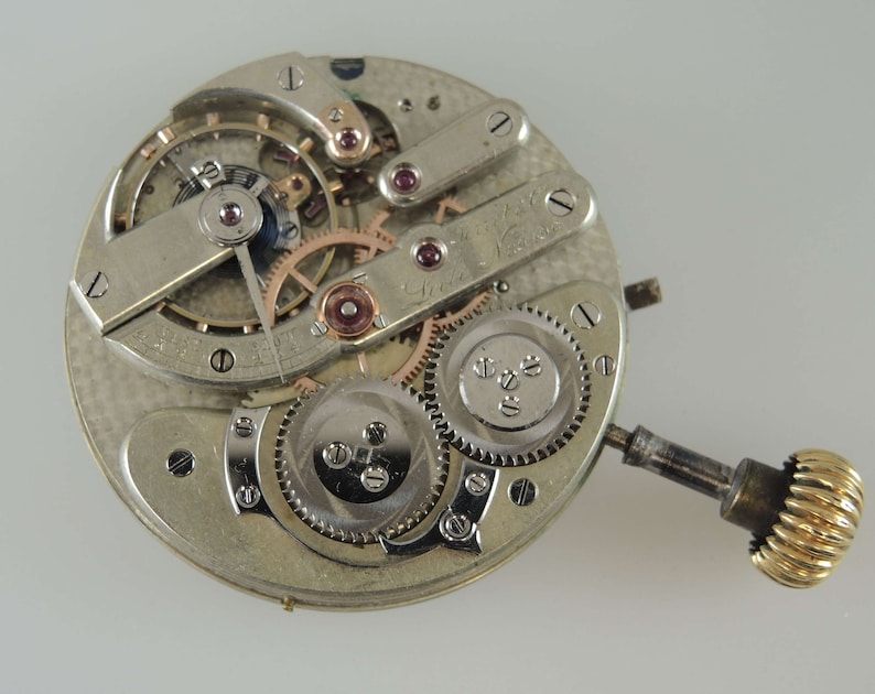 May include: Close-up of a silver pocket watch movement with a gold winding stem. The movement has a number of gears and a balance wheel. The watch is in need of repair.