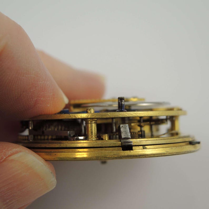 May include: Close-up side view of a gold-toned pocket watch movement with intricate gears and a blue steel screw. The watch is disassembled and the inner workings are visible.