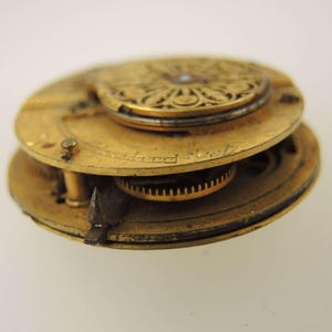 Puede incluir: Primer plano del movimiento de un reloj de bolsillo antiguo. Los componentes de lat&oacute;n incluyen engranajes, placas y una pieza superior decorativa. El mecanismo del reloj est&aacute; parcialmente desmontado, revelando detalles intrincados y el texto "London Fast".