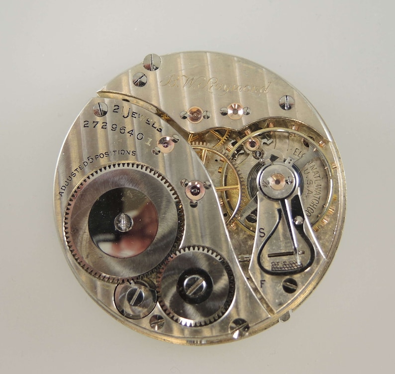 Op de afbeelding: Close-up van een zilveren zakhorloge-uurwerk met de inscriptie "E.L.G. ELGIN NATIONAL WATCH CO. USA" en "21 JEWELS 17 2729640 11 ADJUSTED 5 POSITIONS".