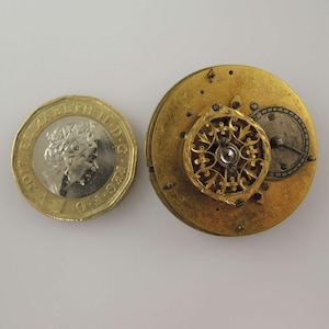 May include: A close-up of a gold-coloured watch movement, with a British one-pound coin for scale. The watch mechanism has a circular design with detailed filigree and a small, silver-coloured dial. The coin features Queen Elizabeth II.