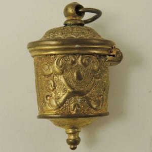 May include: A gold-colored metal thimble with an ornate design and a hinged lid. The thimble has a loop at the top for hanging.