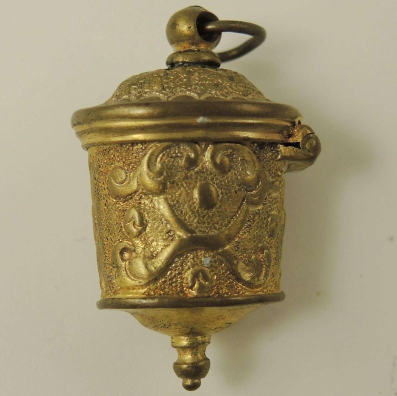 May include: A gold-colored metal thimble with an ornate design and a hinged lid. The thimble has a loop at the top for hanging.