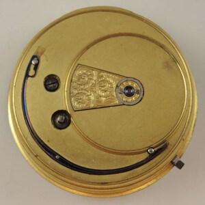 May include: Close-up of a gold-colored pocket watch movement with a decorative engraved plate. The movement has two screws and a winding stem.