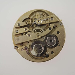 May include: Close-up of a vintage watch movement. The intricate mechanism features numerous gears, screws, and plates in a gold tone. The watch parts are arranged in a circular pattern, showcasing the complexity of the timepiece.