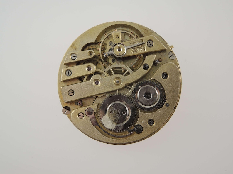 May include: Close-up of a vintage watch movement. The intricate mechanism features numerous gears, screws, and plates in a gold tone. The watch parts are arranged in a circular pattern, showcasing the complexity of the timepiece.