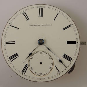May include: A white pocket watch face with black Roman numeral hour markers and black hands. The watch face has a smaller, concentric circle with black markings for seconds. The watch face is labeled "AMERICAN WATCH CO."