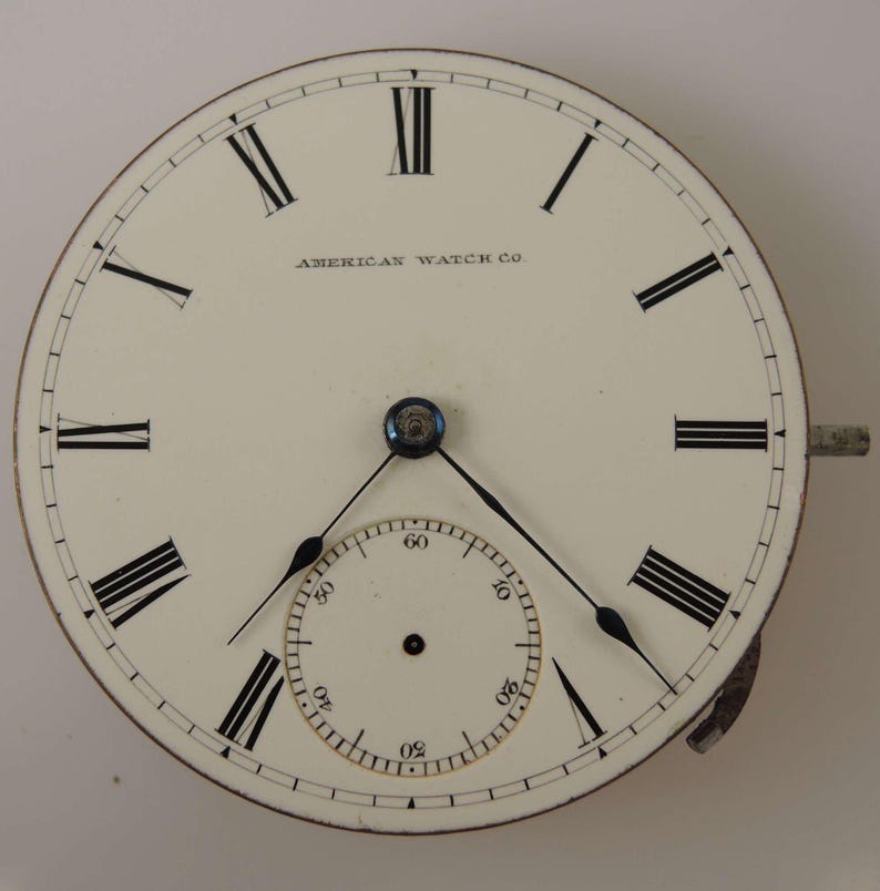 May include: A white pocket watch face with black Roman numeral hour markers and black hands. The watch face has a smaller, concentric circle with black markings for seconds. The watch face is labeled "AMERICAN WATCH CO."