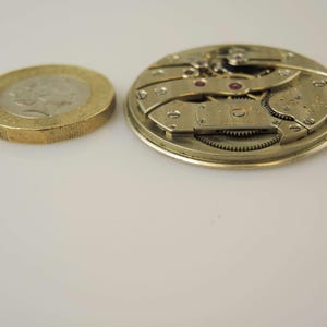 May include: Close-up of a silver-toned watch movement with intricate gears and a small, round, silver-toned plate with the text "Swiss Made" on it.
