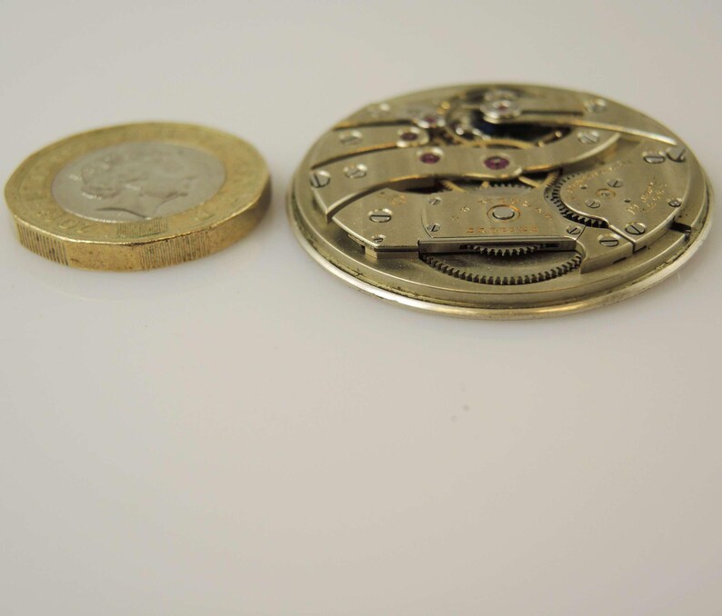 May include: Close-up of a silver-toned watch movement with intricate gears and a small, round, silver-toned plate with the text "Swiss Made" on it.